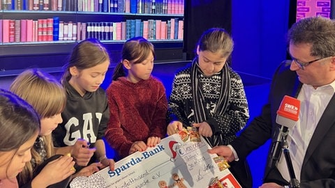 Drei Herzenssache-Kids sitzen auf einer Bühne, daneben sitzt Martin Hettich von der Sparda-Bank Baden-Württemberg (Foto: Herzenssache e.V.)