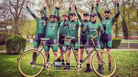 Sieben Männer stehen hinter einem Tandem und heben freudig die Arme.