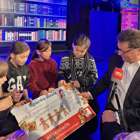 Drei Herzenssache-Kids sitzen auf einer Bühne, daneben sitzt Martin Hettich von der Sparda-Bank Baden-Württemberg (Foto: Herzenssache e.V.)