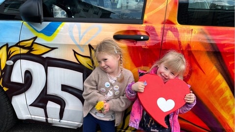 Zwei blonde Mädchen stehen mit Herzenssache-Herz vor dem bunten Herzenssache-Bus (Foto: Herzenssache) Zwei blonde Mädchen stehen mit Herzenssache-Herz vor dem bunten Herzenssache-Bus